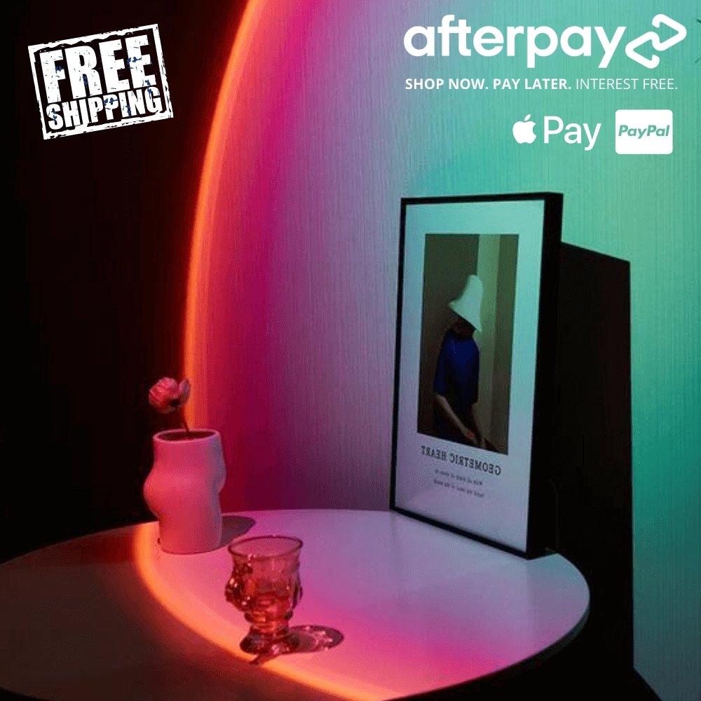 Sunset lamp casting a vibrant gradient of pink, orange, and green light on a wall, illuminating a modern vanity or desk setup with ambient glow.