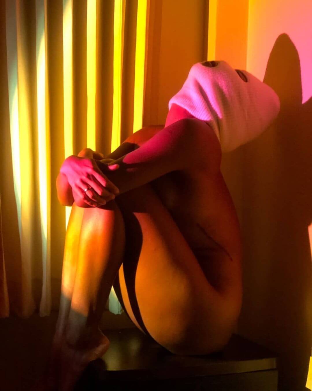 Person sitting with knees up in a warmly lit room, with orange and pink tones from a sunset lamp casting soft shadows on the wall and curtains.