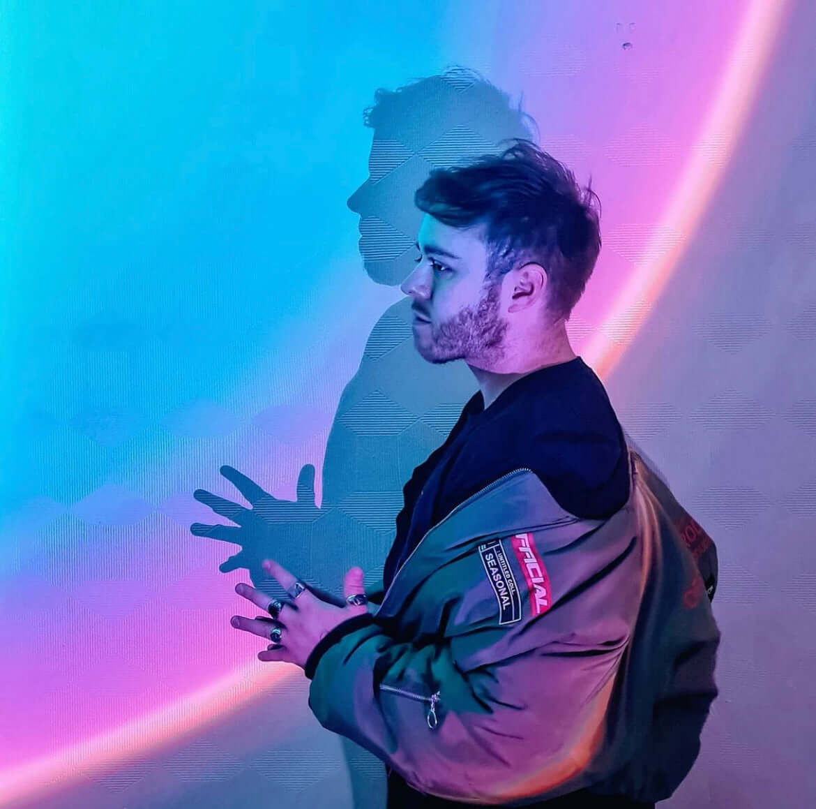Man standing in front of a wall illuminated by a colorful sunset lamp, casting vibrant pink, purple, and blue gradient light with a shadow silhouette effect.